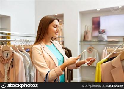 shopping, technology and people concept - happy young woman choosing clothes in mall or clothing store and scanning price tag with smartphone. happy woman using phone app at clothing store