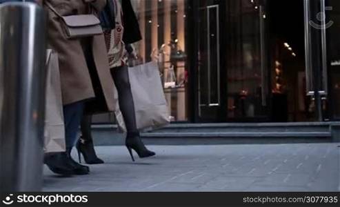 Shopping therapy. Closeup of elegant slim females legs walking down the street and carrying shopping bags after shopping on sunny day over store showcase background. Slow motion. Slender legs of young beautiful girls shopping in the trade center.