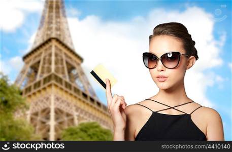 shopping tour, finances, travel, people and luxury concept - beautiful young woman in elegant black sunglasses with credit card over paris eiffel tower background