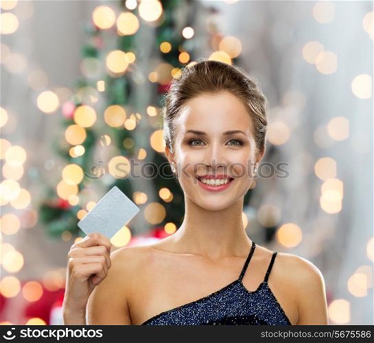 shopping, wealth, holidays and people concept - smiling woman in evening dress holding credit card over christmas tree and lights background
