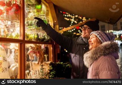 shopping, winter holidays and people concept - happy senior couple at christmas market souvenir shop window on town hall square in tallinn, estonia. happy senior couple hugging at christmas market