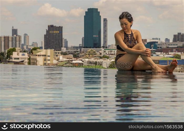 Short haired young woman relax sitting on the edge of roof top outdoor swimming pool and enjoying the city view Bangkok. Summer vacation concept.