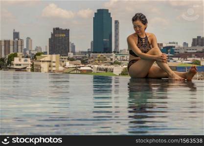 Short haired young woman relax sitting on the edge of roof top outdoor swimming pool and enjoying the city view Bangkok. Summer vacation concept.