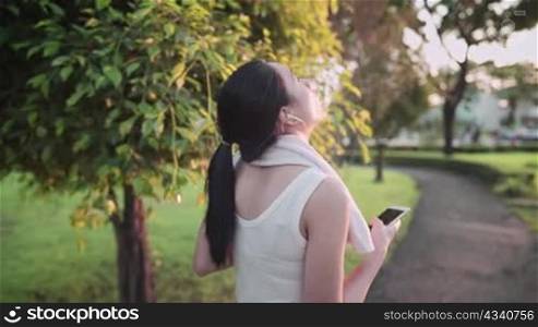 Shot from behind of Young asian woman in exercise clothing wear earphone using smartphone while cooling down at the park during warm sunset hour, relaxing walk wiping sweat from face after work out