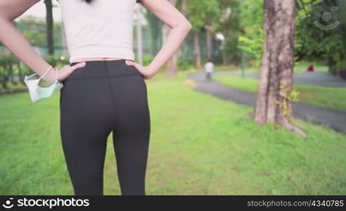 Shot from behind of young woman doing hip waist warm up before running inside the park, green grass field, human body flexibility, doing exercise during Coronavirus pandemic, protective face mask