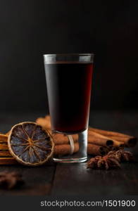 Shot glass of herb liqueur with cinnamon and dry oranges on dark wooden background.