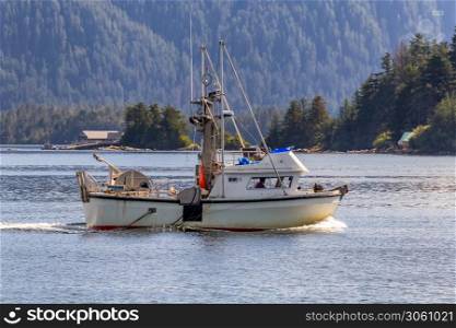 Shot of a commercial fishing boat sailing in a harbour in Sitka, AK. Green forest and a house in the background.