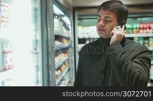 Shot of a man in a food shop. He&acute;s talking on the phone and choosing beverage in the refrigefator.
