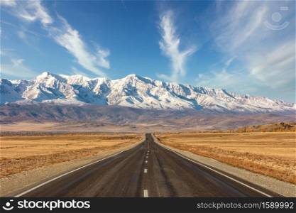 Shot of a straight empty highway leading to the snowy peaks of The Kuray mountain range. Beautiful blue cloudy sky as a background. Altai mountains, Siberia, Russia.. Shot of a straight empty highway leading to the snowy peaks of The Kuray mountain range. Beautiful blue cloudy sky as a background. Altai mountains, Siberia, Russia