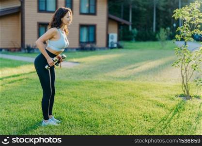 Shot of active sporty woman raises dumbbells and has workout session, works on biceps, dressed in top and leggings, poses on green grass near private house, has athletic body, leads active lifestyle