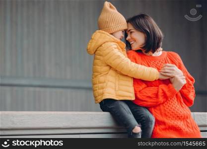 Shot of affectionate young woman in warm red sweater, looks with toothy smile and love at her small daughter, enjoy togetherness, pose against grey background with copy space for your promotion