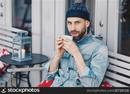 Shot of attractive middle aged male with thick dark beard and mustache, has blue eyes, drinks hot beverage, dressed in jean jacket, looks directly into camera, dreams about something pleasant.