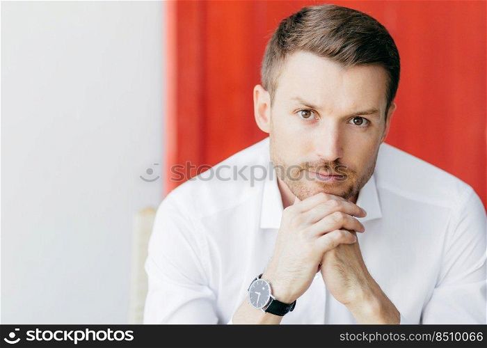 Shot of attractive thoughtful unshaven man holds chin, dressed in elegant white shirt, waits for someone, being prosperous businessman, poses against white and red background with free space aside