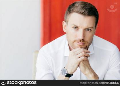 Shot of attractive thoughtful unshaven man holds chin, dressed in elegant white shirt, waits for someone, being prosperous businessman, poses against white and red background with free space aside