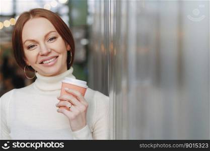 Shot of attractive woman with toothy smile, has makeup, dressed in white turtleneck sweater with collar, holds coffee to go, enjoys spare time, expresses good emotions. People and leisure concept