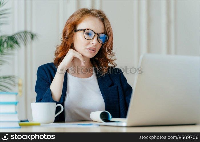 Shot of atttactive businesswoman concentrated in monitor of laptop computer, has serious focused gaze, wears optical glasses for vision correction, watches training video, drinks hot beverage