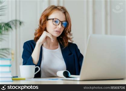 Shot of atttactive businesswoman concentrated in monitor of laptop computer, has serious focused gaze, wears optical glasses for vision correction, watches training video, drinks hot beverage