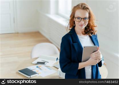 Shot of beautiful red haired young woman checks notification on touch pad, happy to recieve income message, looks confidently at camera, workplace with paper documents and notepad in background.