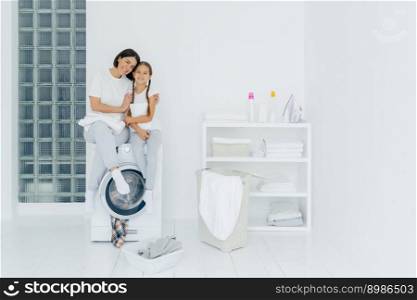 Shot of beautiful woman and his small daughter embrace and smile pleasantly, sit on washing machine, wash linen in laundry room, have friendly relationship, do laundry at home. Housework concept