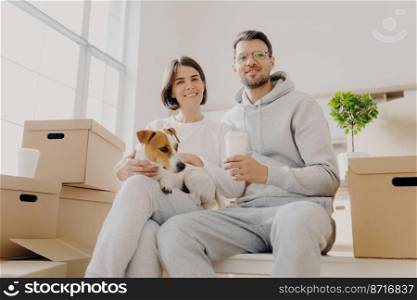 Shot of cheerful husband and wife pose together with pedigree dog, pose in empty room with no furniture, carton boxes with personal belongins, drink takeaway coffee, enjoy togetherness. Moving