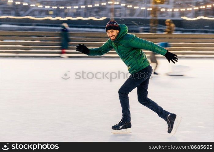 Shot of cheerful male has fun outdoor, skates on ice ring, enjoys this winter activity, moves actively, tries to keep balance, being well trained, looks at camera with happy expression