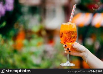 Shot of female`s hand with manicure hold fresh summer drink in wine glass. Orange cold cocktail with ice for you to drink against blurred background