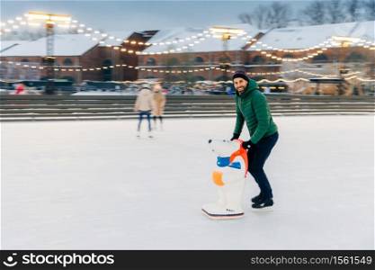 Shot of glad bearded male spends time outdoor on fresh air, enjoys frosty winter weather, goes skating on ice ring, uses skae aid as afraid to fall down. Handsome man has fun on skating ring
