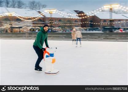 Shot of glad bearded male spends time outdoor on fresh air, enjoys frosty winter weather, goes skating on ice ring, uses skae aid as afraid to fall down. Handsome man has fun on skating ring