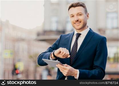 Shot of handsome male entrepreneur comes on meeting with business people, looks at wristwatch, uses touchpad for searching necessary information in internet, wears elegant formal suit and tie