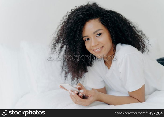 Shot of lovely curly woman with dark skin, tender smile on face, lies on white bed, wears t shirt, has relax with modern technologies, poses in bedroom, reads text message, uses free internet