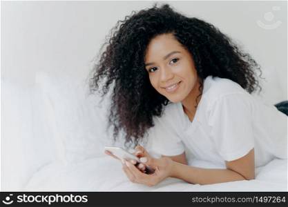 Shot of lovely curly woman with dark skin, tender smile on face, lies on white bed, wears t shirt, has relax with modern technologies, poses in bedroom, reads text message, uses free internet