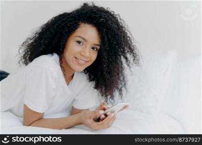 Shot of lovely curly woman with dark skin, tender smile on face, lies on white bed, wears t shirt, has relax with modern technologies, poses in bedroom, reads text message, uses free internet