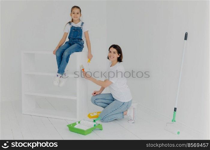 Shot of lovely mother and small hard working daughter pose in empty room, paint furniture in white color, use paintbrushes, happy to finish work, being busy during weekend. People, repair, improvement
