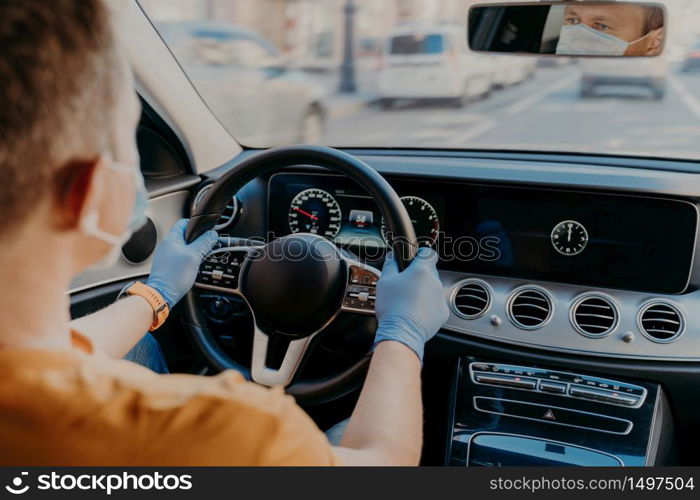 Shot of man driver wears protective medical mask and rubber gloves, poses in car, cares about health during coronavirus. Safety in transport. Health care