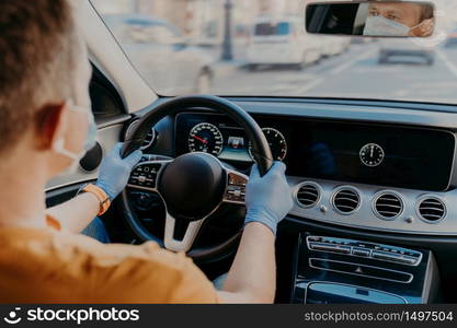 Shot of man driver wears protective medical mask and rubber gloves, poses in car, cares about health during coronavirus. Safety in transport. Health care