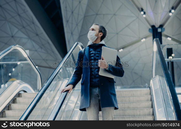 Shot of man tourist poses on escalator in airport, arrives home from abroad during virus outbreak, wears protective medical mask. Evacuated passenger. Infectious disease. Public safety concept