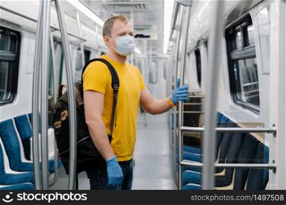 Shot of man wears disposable medical mask during coronavirus outbreak, keeps safety, poses in empty carriage of underground, carries rucksack, commutes to work. Covid-19 and health care concept