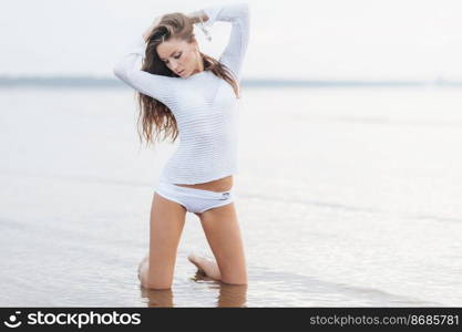 Shot of pleasant looking female in white summer clothing, has luxurious long dark hair, stands on knees at sand near sea, poses for women s magazine, looks down. Woman has summer vacations