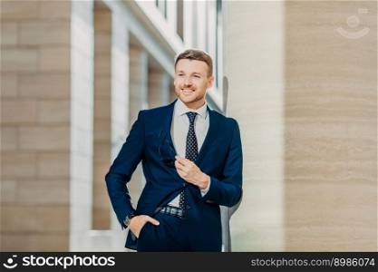Shot of pleased successful male manager with bristle, wears formal clothes, looks confidently and happily aside, stands in office building, being sure in success. People, career and work concept
