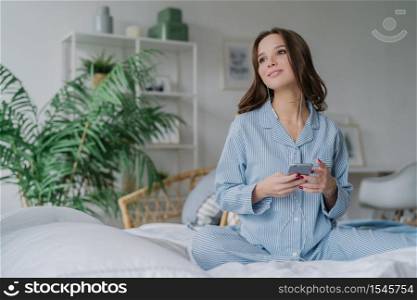 Shot of pretty brunette student learns language via mobile app, listens audio with earphones, has free time, dressed in casual outfit, poses on comfortable white bed, has thoughtful look aside