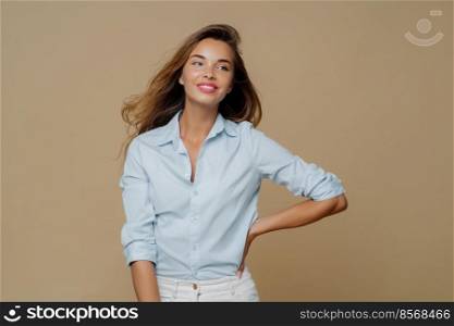 Shot of pretty cheerful European woman keeps one hand on waist, looks aside, has charming smile wears shirt and trousers, has slim perfect figure, enjoys shopping day, isolated over brown background