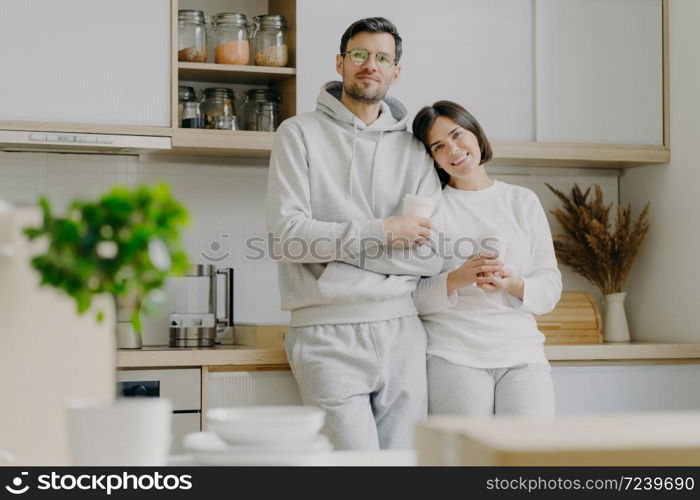 Shot of relaxed family couple stand in kitchen, drink takeaway coffee, wear casual outfit, pose in kitchen interior, bought new apartment for living. People, real estate and mortgage concept