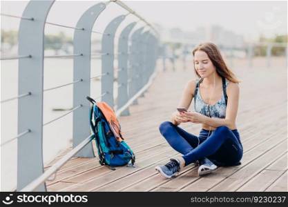 Shot of satisfied lovely female likes sport and active lifestyle, wears top and leggings, reads positive news in internet on cellular, rests after covering long distance on foot, listens music