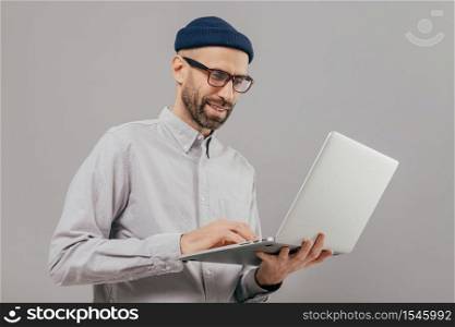 Shot of satisfied male with thick beard and mustache, holds laptop computer, transfers money, uses online banking service, sends files, connected to wireless internet, models over grey background