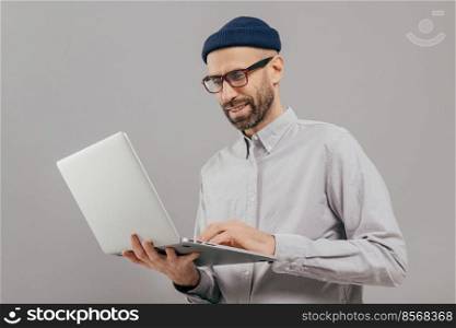 Shot of satisfied male with thick beard and mustache, holds laptop computer, transfers money, uses online banking service, sends files, connected to wireless internet, models over grey background