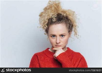 Shot of serious blonde female keeps hand under chin, looks seriously at camera, isolated over white concrete wall. Serious student listens important information for exam, poses in studio alone