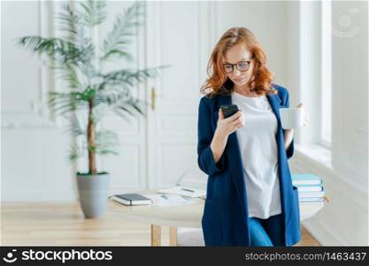 Shot of serious female entrepreneur focused in mobile phone, makes payment via banking application, drinks aromatic coffee, recieves notification, wears in elegant formal suit, poses indoor.