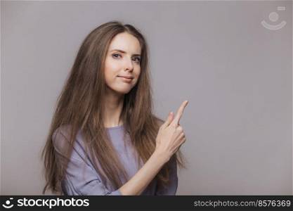 Shot of serious pleasant looking brunette female with blue eyes, dressed casually, points with index finger at blank copy space, isolated over grey background. Woman advertizes new product indoor