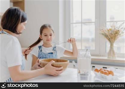 Shot of small kid helper whisks cream with beater, learns how to bake, listens useful advice from mother who holds her pony tail, sit at white kitchen table together. Family cooking together
