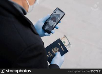 Shot of unrecognizable man follow rules of social distancing, makes video call, holds mobile phone, passport with money wears protective gloves walks outdoor at street. Avoid contact with other people